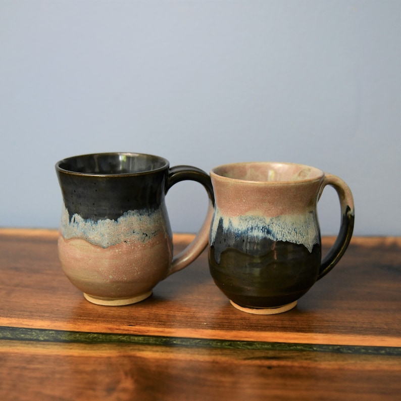 May include: Two handmade ceramic mugs with a unique glaze. One mug is dark with a blue and tan band, the other is brown with a blue and tan band. Both mugs have a curved handle and sit on a wooden surface.