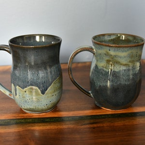 May include: Two handmade ceramic mugs with a dark blue and green glaze. Each mug has a large handle and a unique color gradient, transitioning from dark blue to a lighter green. The mugs are displayed on a wooden surface.