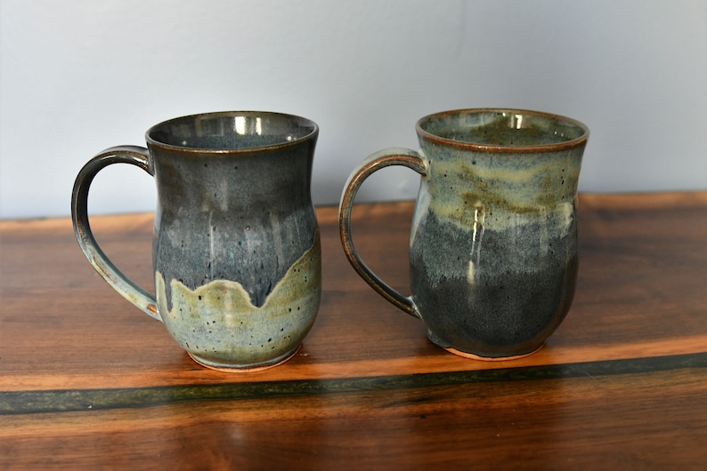 May include: Two handmade ceramic mugs with a dark blue and green glaze. Each mug has a large handle and a unique color gradient, transitioning from dark blue to a lighter green. The mugs are displayed on a wooden surface.