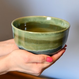 May include: A handmade ceramic bowl with a glossy finish. The bowl has a gradient of green and blue tones, with a darker band near the base. The bowl is held in two hands.