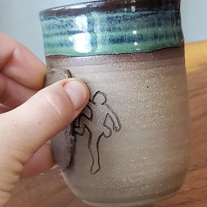 May include: A handmade ceramic mug with a climbing figure etched on the side. The mug has a speckled, light-coloured body with a dark brown rim and a band of teal green. A small, textured handle is attached to the side.