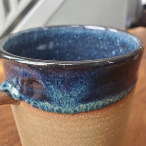 May include: Close-up of a handmade ceramic mug. The mug features a speckled, light brown base and a dark blue interior and rim. The rim has a dark, glossy finish. The handle is a matching brown color. The mug is designed for hot beverages.