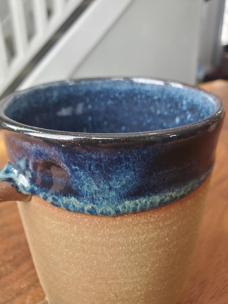 May include: Close-up of a handmade ceramic mug. The mug features a speckled, light brown base and a dark blue interior and rim. The rim has a dark, glossy finish. The handle is a matching brown color. The mug is designed for hot beverages.