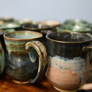 May include: A collection of handmade ceramic mugs with unique color gradients. The mugs feature a dark brown to light pink and blue glaze, with visible brushstrokes and a glossy finish. Each mug has a rounded handle and a rustic, artisanal aesthetic.
