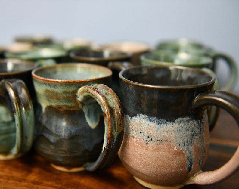 May include: A collection of handmade ceramic mugs with unique color gradients. The mugs feature a variety of earthy tones, including greens, blues, and browns, with a glossy finish. Each mug has a sturdy handle and a wide opening, perfect for coffee or tea.