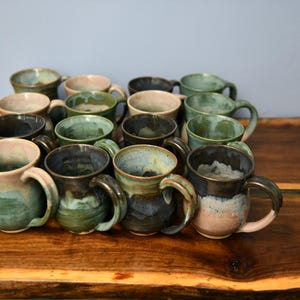 May include: A collection of handmade ceramic mugs in various colors, including green, blue, and brown. Each mug has a unique glaze and a sturdy handle. The mugs are arranged on a wooden surface.