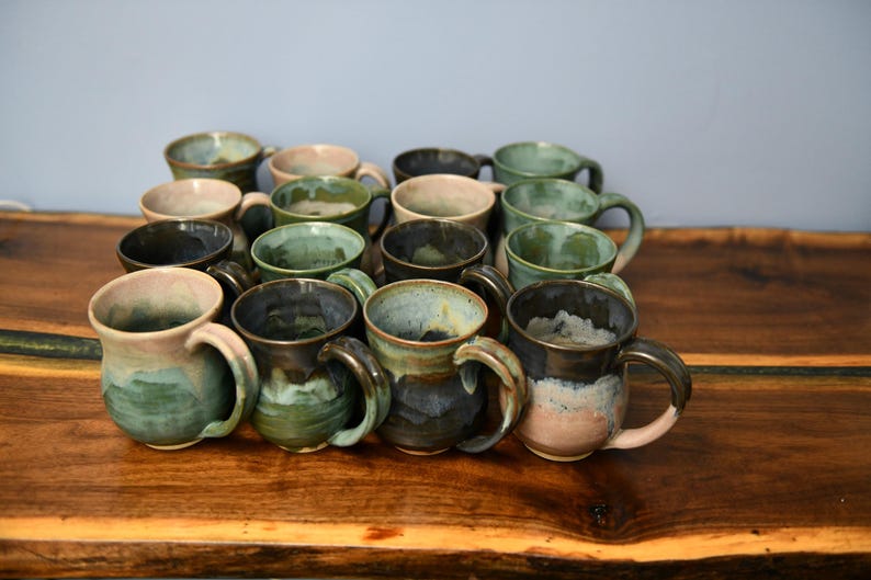 May include: A collection of handmade ceramic mugs in various colors, including green, blue, and brown. Each mug has a unique glaze and a sturdy handle. The mugs are arranged on a wooden surface.