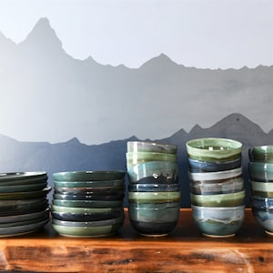 May include: A selection of handmade ceramic bowls and plates in shades of green, blue, and grey. The tableware is stacked on a wooden surface against a backdrop of a mountain range mural. The bowls and plates have a rustic, artisanal aesthetic.