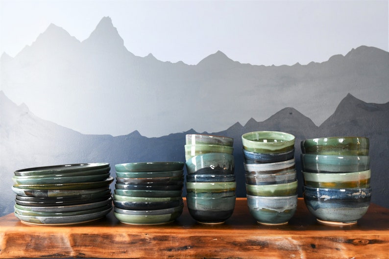 May include: A selection of handmade ceramic bowls and plates in shades of green, blue, and grey. The tableware is stacked on a wooden surface against a backdrop of a mountain range mural. The bowls and plates have a rustic, artisanal aesthetic.