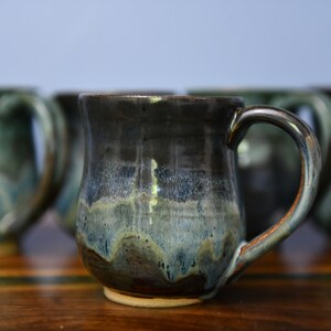 May include: A handmade ceramic mug with a dark, glossy glaze that transitions from black to blue and green. The mug has a rounded body, a sturdy handle, and a light-colored base. Other mugs are visible in the background.