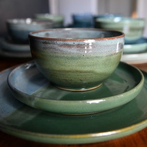 Può includere: Un set di stoviglie in ceramica fatte a mano nei toni del verde e del blu. Il set comprende ciotole e piatti, con finitura lucida. Le ciotole sono impilate sui piatti. I pezzi in ceramica hanno un aspetto rustico e artigianale, perfetti per servire i pasti.
