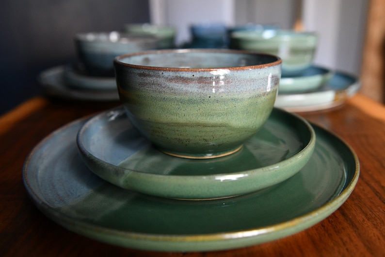 May include: Handmade ceramic tableware set in green and blue hues. The set includes bowls and plates with a glossy finish. The bowls are stacked on the plates. The ceramic pieces have a rustic, artisanal appearance, ideal for serving meals.