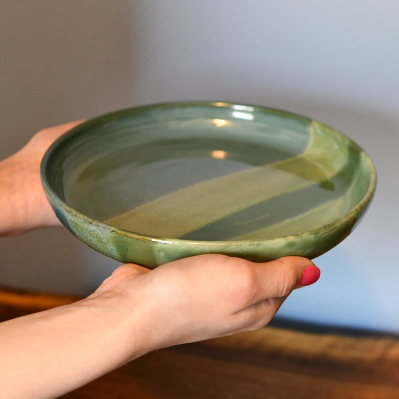 May include: A handmade ceramic serving bowl with a two-tone green glaze. The bowl has a wide, shallow design and a slightly textured rim. The glaze transitions from a darker green to a lighter green, creating a subtle diagonal pattern.
