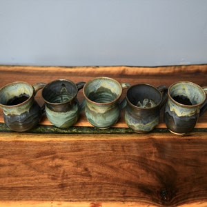 May include: Five handmade ceramic mugs with a unique, textured design. The mugs feature a blend of blue, green, and brown glazes, creating a rustic and artistic appearance. Each mug has a handle and a slightly irregular shape, showcasing the handcrafted nature of the product.