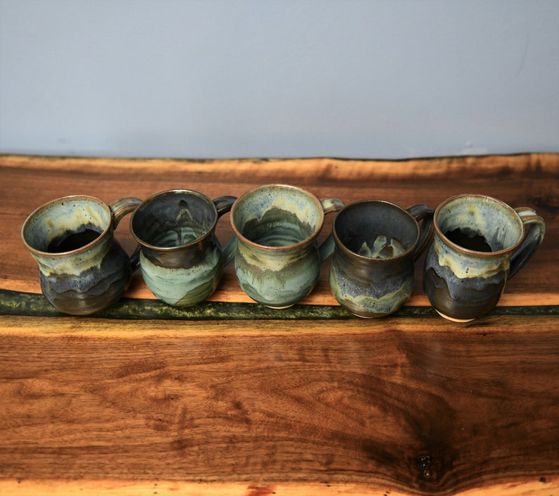 May include: Five handmade ceramic mugs with a unique, textured design. The mugs feature a blend of blue, green, and brown glazes, creating a rustic and artistic appearance. Each mug has a handle and a slightly irregular shape, showcasing the handcrafted nature of the product.