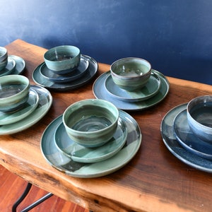 May include: A selection of handmade ceramic tableware sets. Each set comprises a bowl and two plates in shades of blue, green, and grey. The dishware is displayed on a wooden table, highlighting the unique shapes and colour variations.
