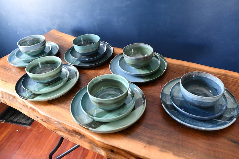 May include: A selection of handmade ceramic tableware sets. Each set comprises a bowl and two plates in shades of blue, green, and grey. The dishware is displayed on a wooden table, highlighting the unique shapes and colour variations.