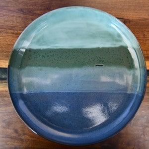 May include: A round ceramic plate with a gradient of colours. The top is light teal, transitioning to a darker teal, then a deep green, and finally a dark blue at the bottom. The plate is on a wooden surface.