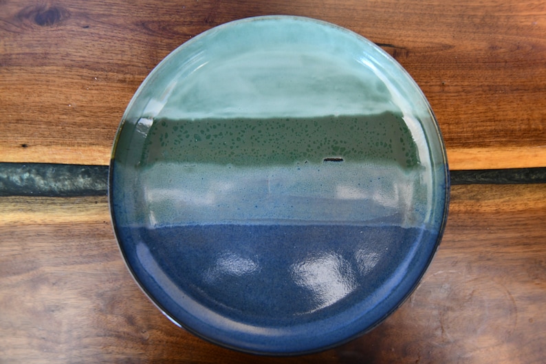 May include: A round ceramic plate with a gradient of colours. The top is light teal, transitioning to a darker teal, then a deep green, and finally a dark blue at the bottom. The plate is on a wooden surface.