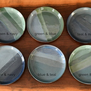 May include: Six ceramic plates with a two-tone design in shades of green, blue, and navy. Each plate features a different colour combination, with text labels such as "green & navy" and "blue & teal". The plates are arranged on a wooden surface.