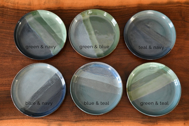May include: Six ceramic plates with a two-tone design in shades of green, blue, and navy. Each plate features a different colour combination, with text labels such as "green & navy" and "blue & teal". The plates are arranged on a wooden surface.