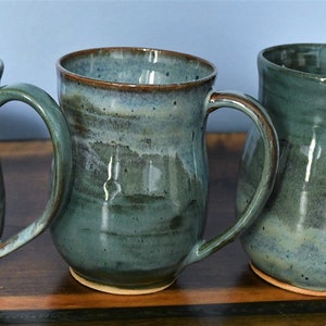 May include: Three handmade ceramic mugs with a teal glaze and brown rims. Each mug has a large handle and a unique shape, showcasing the artistry of the pottery. The mugs are displayed on a wooden surface.