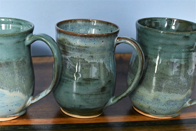 May include: Three handmade ceramic mugs with a teal glaze and brown rims. Each mug has a large handle and a unique shape, showcasing the artistry of the pottery. The mugs are displayed on a wooden surface.