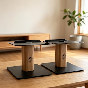 Könnte beinhalten: Zwei Lautsprecherständer mit flachen, quadratischen Bodenplatten und Oberteilen aus schwarzem Metall, verbunden durch eine vertikale Säule aus hellem Holz. Jede Holzsäule zeigt einen schwarzen Aufdruck eines Violinschlüssels mit Verzierungen.