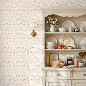 May include: A cream-colored hutch with open shelving displays white ceramic plates, bowls, and jars. A woven basket holds bread, while a vase of flowers sits on the top shelf. A wall-mounted lamp with a pleated shade is attached to floral wallpaper.