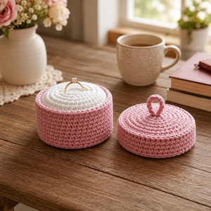 May include: A round, pink crocheted jewelry box with a white crocheted insert holding a gold ring. Beside it sits a matching pink crocheted lid with a small loop handle on top. The items are crafted from textured yarn in a circular, dense stitch pattern.