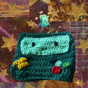 May include: A dark teal crochet pouch featuring a light mint green rectangular face with two black eyes and a small smile. The front includes a yellow star-shaped button, a red circular button, and two smaller green circular buttons.