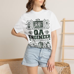 Puede incluir: Una camiseta blanca de manga corta con un estampado gráfico en verde oscuro. El diseño incluye patrones de circuitos, iconos de un bicho, una lista de verificación y el texto "QA ENGINEER" seguido de la frase "I find the bugs before users do."