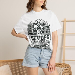 May include: A white short-sleeve t-shirt featuring a black graphic print of a DevOps engineer theme. The design includes icons of servers, a cloud, a gear, and the text "DEVOPS ENGINEER" with the phrase "Automate. Deploy. Repeat." below it.