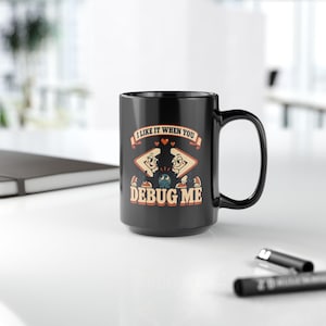 May include: A black ceramic mug featuring a graphic of two cartoon sandwich halves looking at a small blue bug. The text reads I LIKE IT WHEN YOU DEBUG ME in a retro font. The design includes small red hearts between the sandwich characters.