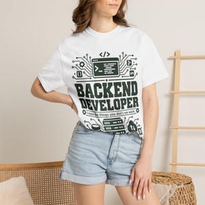 Może przedstawiać: Biały t-shirt z krótkim rękawem z ciemnozielonym nadrukiem graficznym. Wzór zawiera okno kodu komputerowego, linie obwodów drukowanych oraz tekst "BACKEND DEVELOPER", a pod nim "I make the things you don't see work."