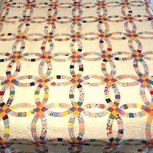 May include: A double wedding ring quilt featuring a cream-colored background with intricate interlocking rings composed of various multi-colored fabric patches. The design includes small solid orange and purple square connectors at each intersection.