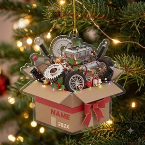 May include: A flat, die-cut Christmas ornament shaped like an open cardboard box filled with automotive parts including a silver engine, gears, and tires. The box features a red ribbon, the text NAME, and the year 202X printed on the front side.