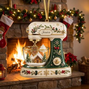 May include: A cream and dark green stand mixer ornament featuring gold accents, holly leaf patterns, and snowflakes. The glass mixing bowl contains a miniature snow-covered house and pine trees. The name Jessica is printed on the front.