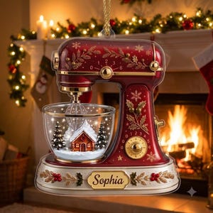May include: A red and white stand mixer Christmas ornament featuring gold accents, snowflake and holly patterns, and a clear bowl containing a miniature winter scene with a house and pine trees. The base displays the name Sophia in gold script.