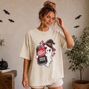 May include: A beige short-sleeved t-shirt featuring a graphic of a white ghost wearing a black witch hat and holding a black Dr. Pepper Zero Sugar soda can. The design includes small red star accents surrounding the ghost and the soda can.