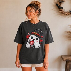 May include: A charcoal gray short-sleeve t-shirt featuring a graphic of a white ghost wearing a black witch hat with a red band. The ghost is holding a red can labeled Dr Pepper Zero Sugar. Small red hearts and four-pointed stars surround the ghost.