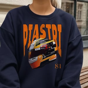May include: A navy blue crewneck sweatshirt featuring the orange text PIASTRI above a graphic of a racing helmet with yellow, black, and white details. The number 81 is printed in orange on the lower right side of the graphic design.