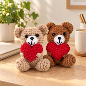 May include: Two small, handmade crochet teddy bears sitting side by side. The bear on the left is light tan, and the bear on the right is medium brown. Both bears have black plastic eyes, white muzzles, and are holding bright red crochet hearts.