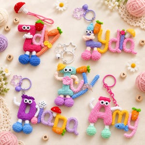 May include: Five handmade crochet keychains featuring names spelled out in colorful yarn letters. Each lettered piece includes attached googly eyes, small crocheted limbs, and decorative elements like tiny carrots, mushrooms, flowers, and bows, all attached to metal key rings.