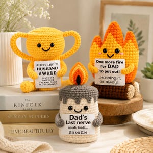 May include: Three crocheted figures: a yellow trophy holding a sign reading World's Greatest Husband Award, an orange flame holding a sign reading One more fire for DAD to put out, and a gray candle holding a sign reading Dad's Last nerve.