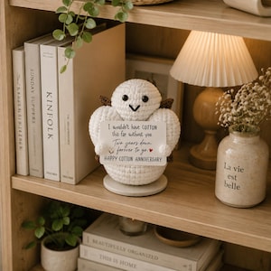 May include: A white crocheted figure with black eyes and a small brown accent, holding a rectangular sign that reads: I wouldn't have COTTON this far without you. Two years down. forever to go. HAPPY COTTON ANNIVERSARY. The figure sits on a round base.
