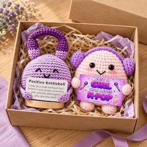 May include: A crochet kettlebell in purple and a crochet character in light pink with purple headphones, both nestled in a gift box. The kettlebell holds a card reading Positive Kettlebell, and the character holds a card reading Just a Girl Who Loves K-Pop.