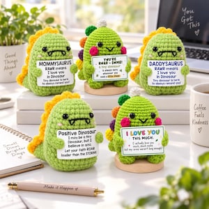 May include: Five crocheted green dinosaur figurines, each holding a white sign with unique text. The dinosaurs feature yellow or multicolored spikes, black eyes, and pink cheeks. They are displayed on small wooden bases or white surfaces.