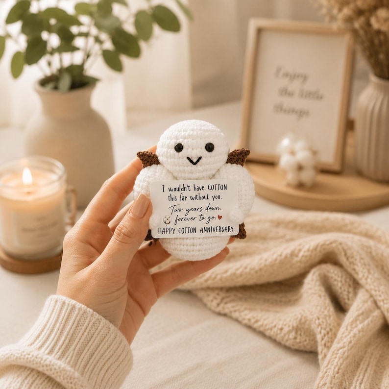 May include: A small, round, white crocheted cotton boll figure with dark brown accents, black eyes, and a V-shaped mouth. It holds a white rectangular card with the text: I wouldn't have COTTON this far without you. Two years down, forever to go. HAPPY COTTON ANNIVERSARY.
