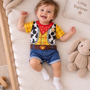 Könnte beinhalten: Ein kurzärmeliger Baby-Strampler mit gelbem Karomuster, einem roten Bandana-Kragen mit weißen Sternen, einer schwarz-weißen Kuhfell-Weste, einem braunen Gürtel mit goldener Schnalle und einer blau aussehenden Jeans-Shorts mit weißen Socken.